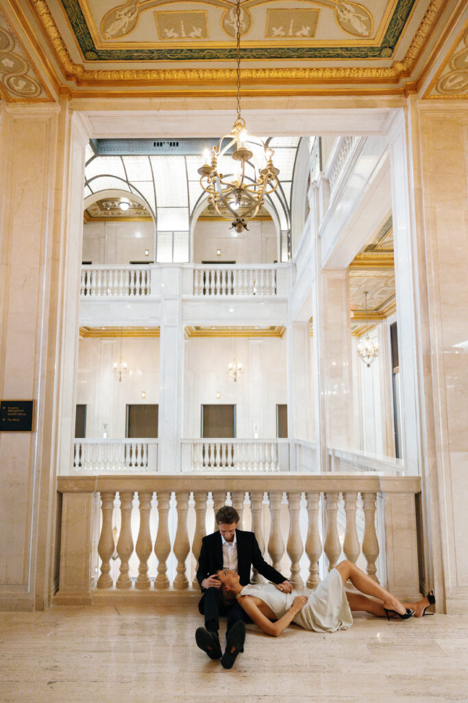 Engaged couple seated on marble floor beneath chandelier in the grand interior of Book Tower Detroit during their Detroit Book Tower engagement session