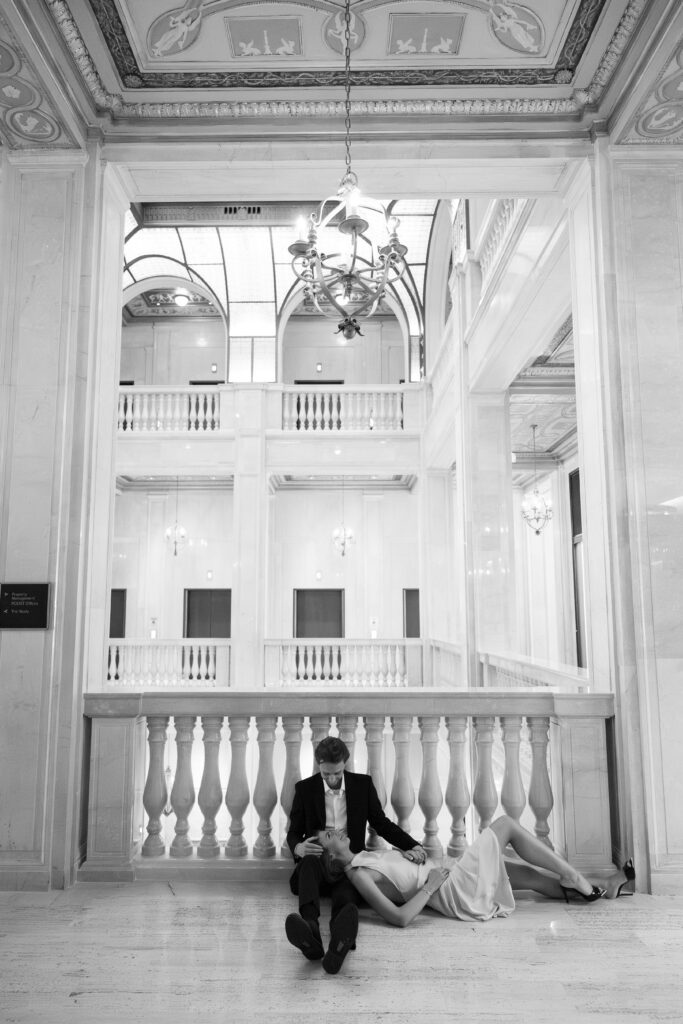 Black and white photo of engaged couple seated on the marble floor in the grand interior of Book Tower during their Detroit Book Tower engagement session