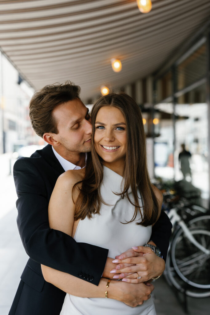 Groom-to-be hugging his smiling fiancée under the striped overhang at Shinola Hotel in downtown Detroit during their Detroit Book Tower engagement session