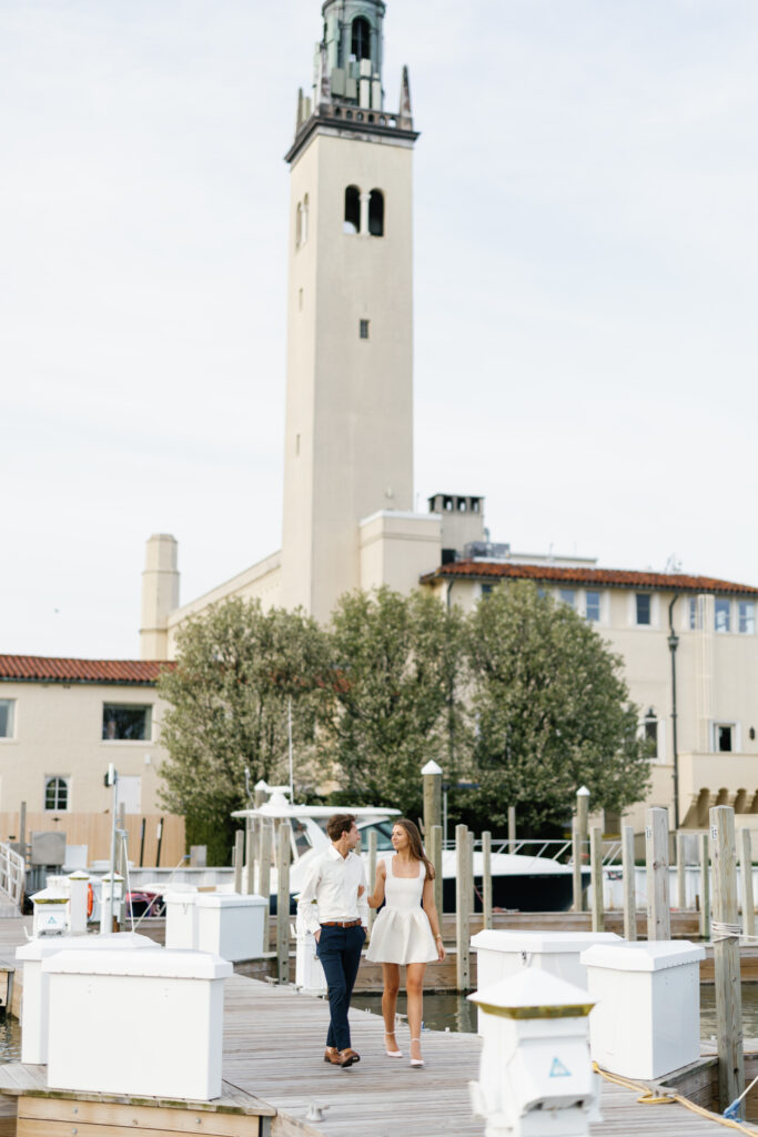 Engaged couple walking hand-in-hand on the dock at Grosse Pointe Yacht Club with the iconic tower in the background during their Detroit Book Tower engagement session