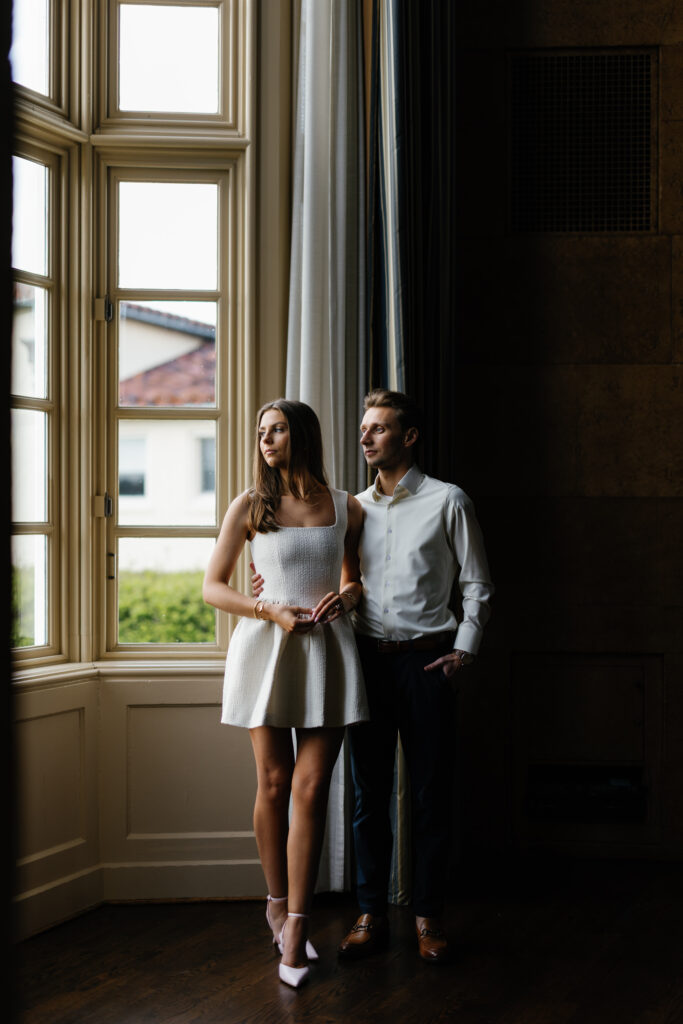 Engaged couple standing by a large window inside Grosse Pointe Yacht Club, softly lit during their Detroit Book Tower engagement session