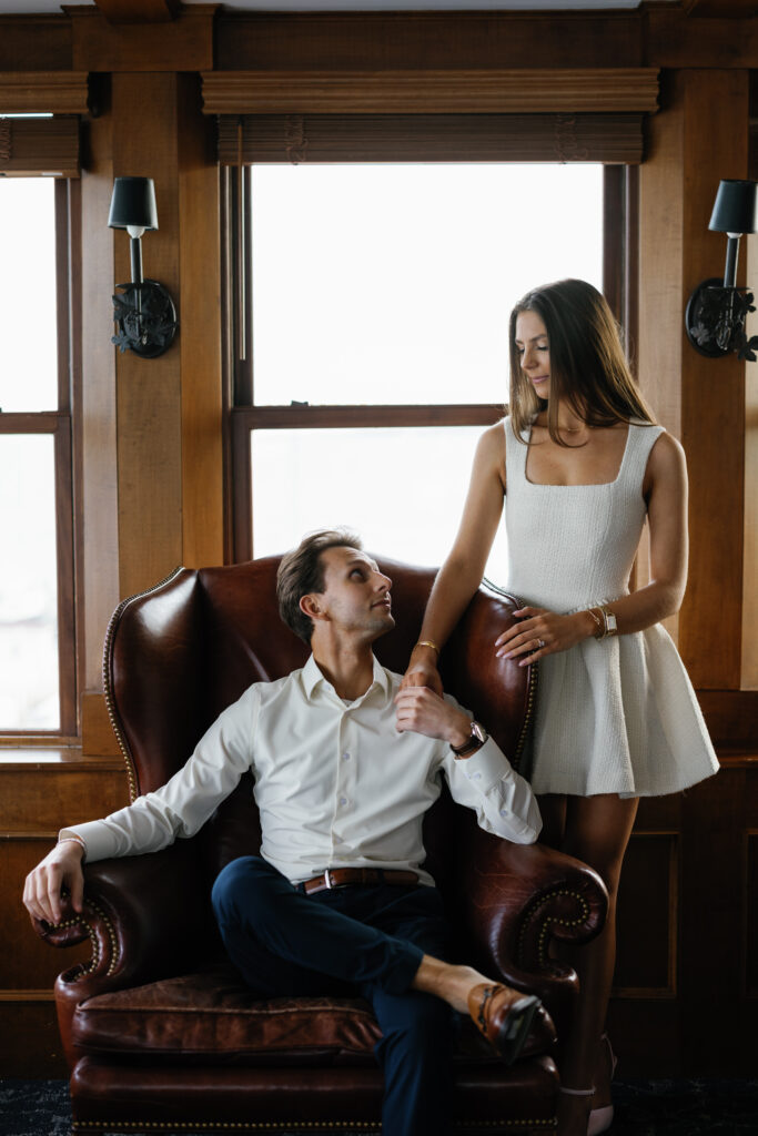 Engaged couple posing in a leather armchair at Grosse Pointe Yacht Club during their Detroit Book Tower engagement session, surrounded by dark wood paneling
