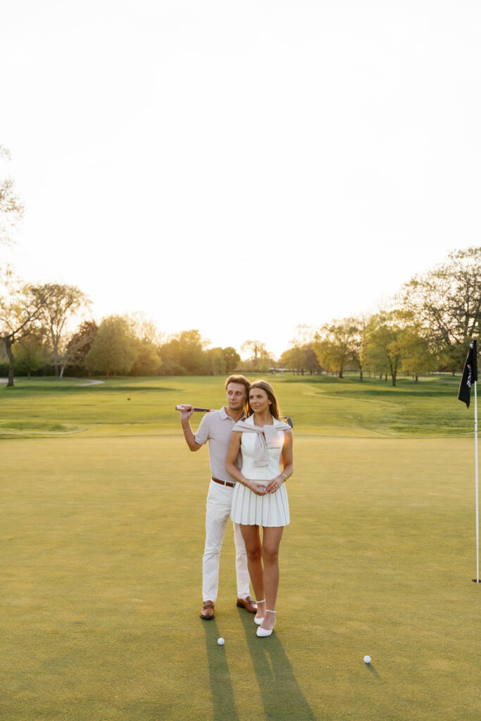 Engaged couple on the green at Lochmoor Golf Club during sunset, dressed in golf attire for their Detroit Book Tower engagement session