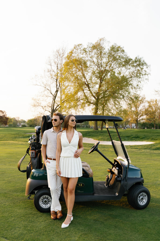 Engaged couple posing in golf-inspired outfits next to a green golf cart at Lochmoor Golf Club during their Detroit Book Tower engagement session