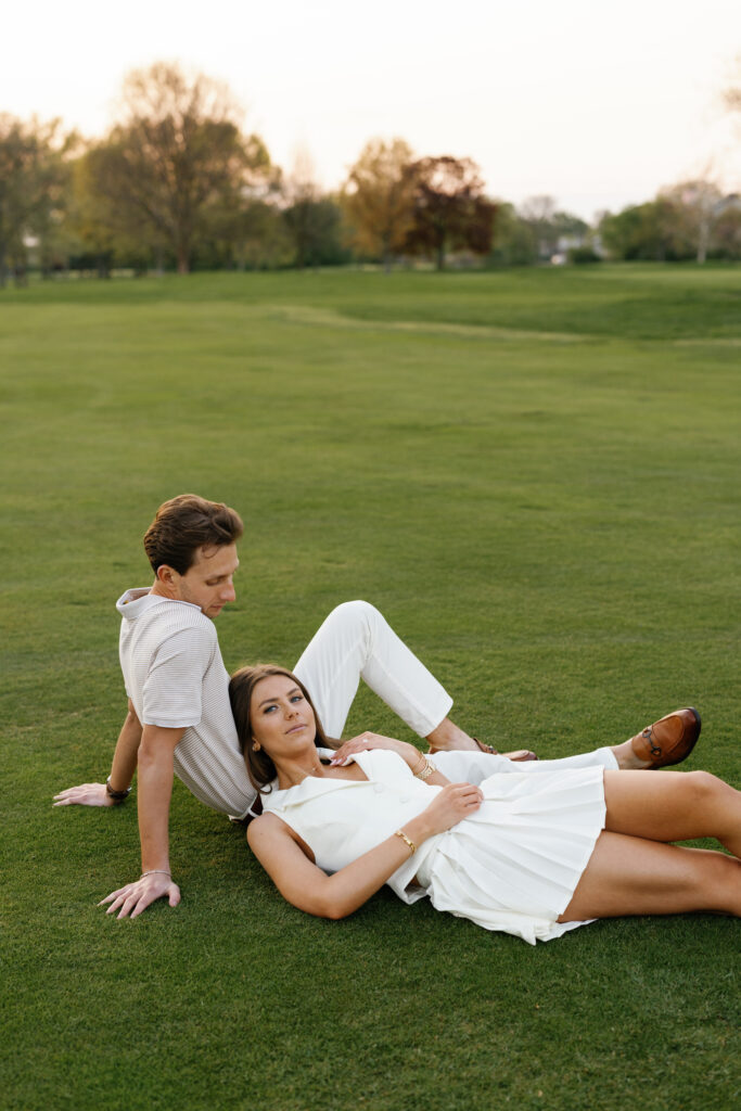 Engaged couple relaxing on the fairway at Lochmoor Golf Club during their Detroit Book Tower engagement session, dressed in white golf-inspired outfits