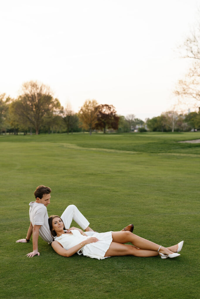 Engaged couple lounging on the green at Lochmoor Golf Club during their Detroit Book Tower engagement session, dressed in coordinated white outfits