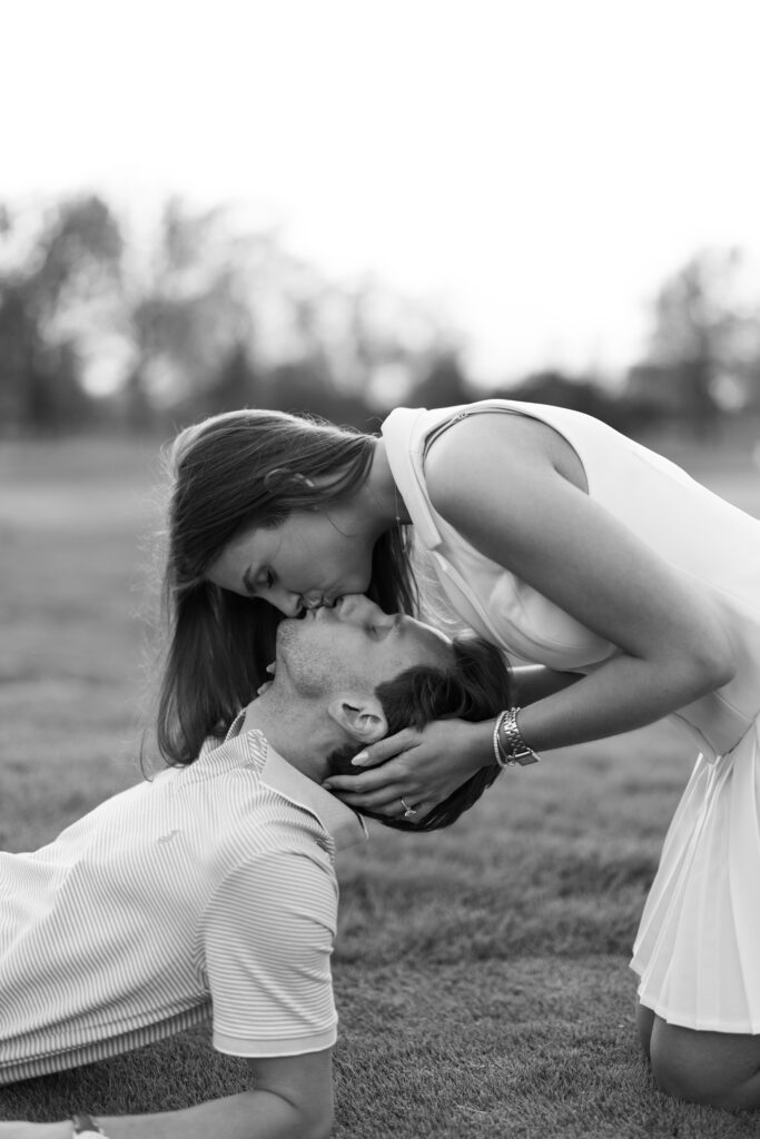 Black and white photo of bride-to-be gently kissing her fiancé’s forehead on the grass at Lochmoor Golf Club during their Detroit Book Tower engagement session