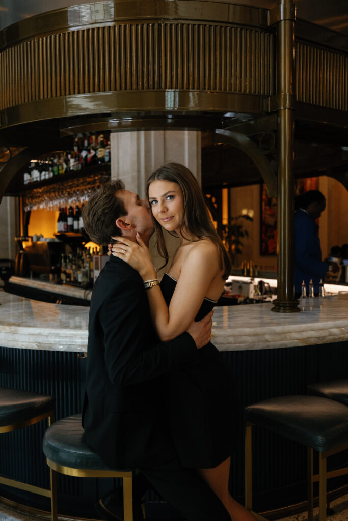 Bride-to-be sitting on her fiancé’s lap at the marble bar inside Book Tower during their Detroit Book Tower engagement session, both dressed in black