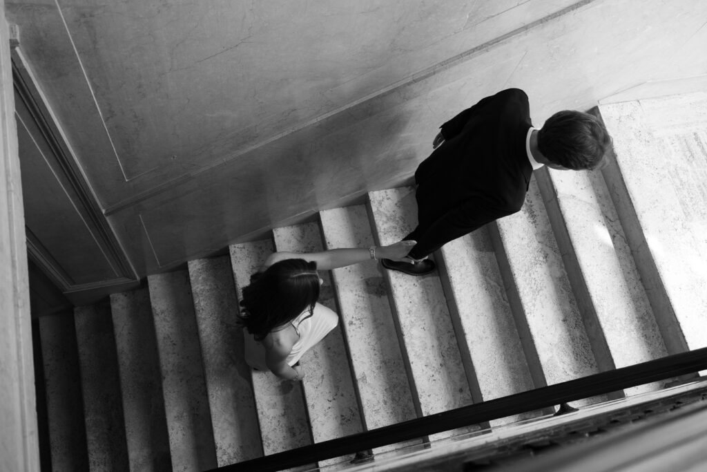 Black and white photo of bride-to-be reaching for her fiancé’s hand as they walk up marble stairs at Book Tower during their Detroit Book Tower engagement session