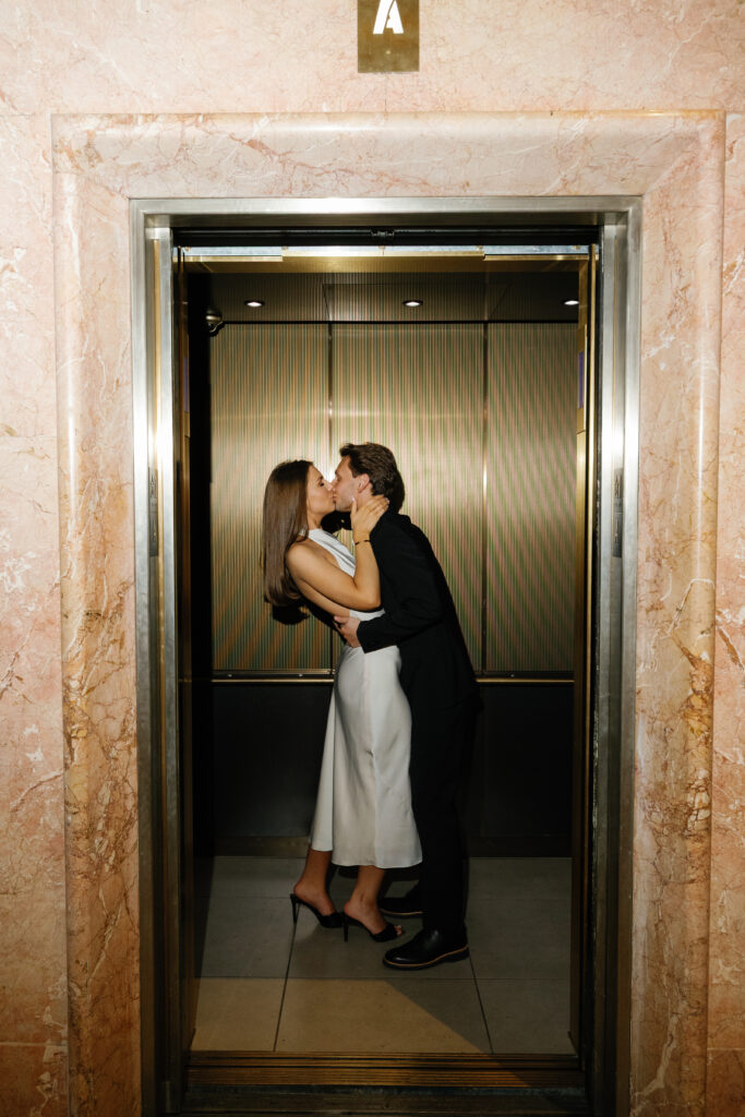 Engaged couple kissing inside gold elevator at Book Tower during their Detroit Book Tower engagement session, framed by pink marble walls
