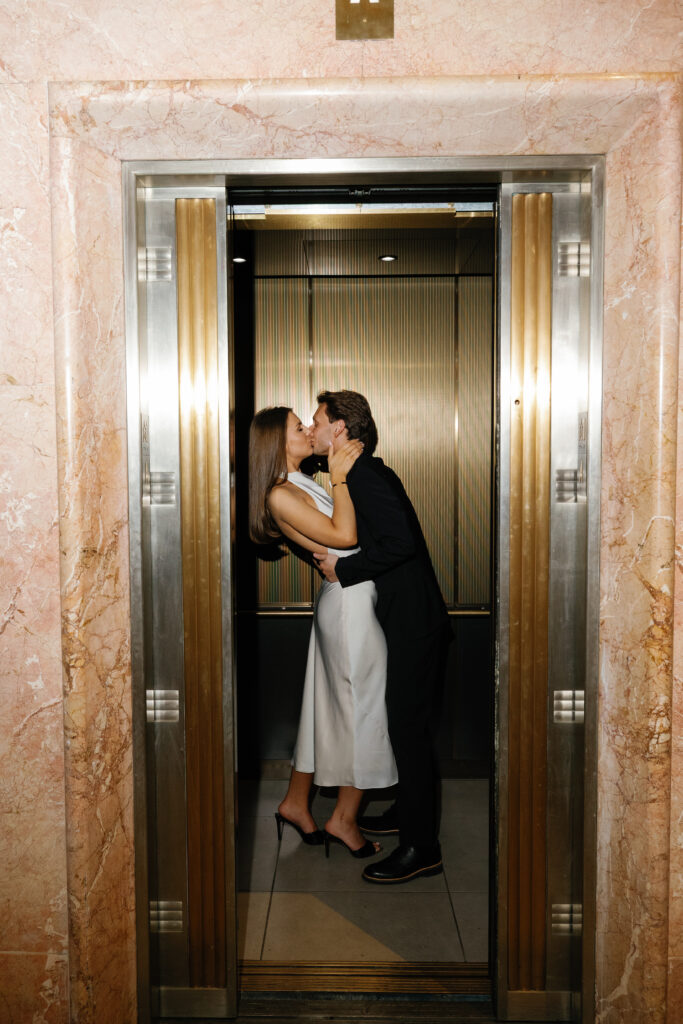 Engaged couple kissing in the elevator doorway framed by pink marble and gold details at Book Tower during their Detroit Book Tower engagement session