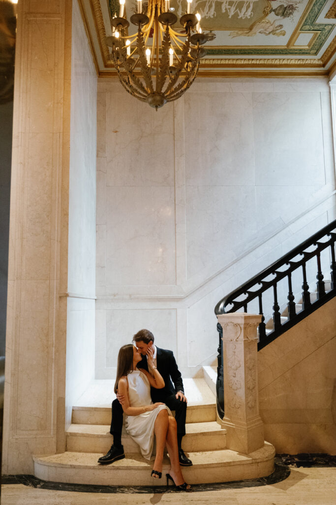 Engaged couple sharing a kiss on marble steps beneath chandelier at Book Tower Detroit during their Detroit Book Tower engagement session