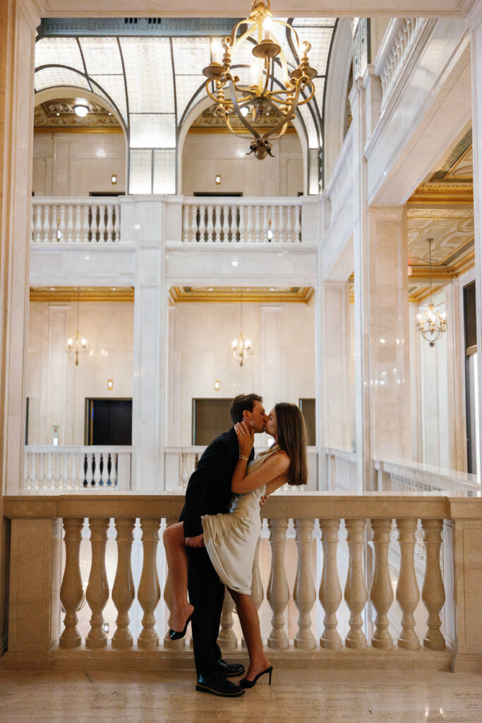 Engaged couple kissing in the grand marble hall of Book Tower, framed by chandeliers and arched windows during their Detroit Book Tower engagement session
