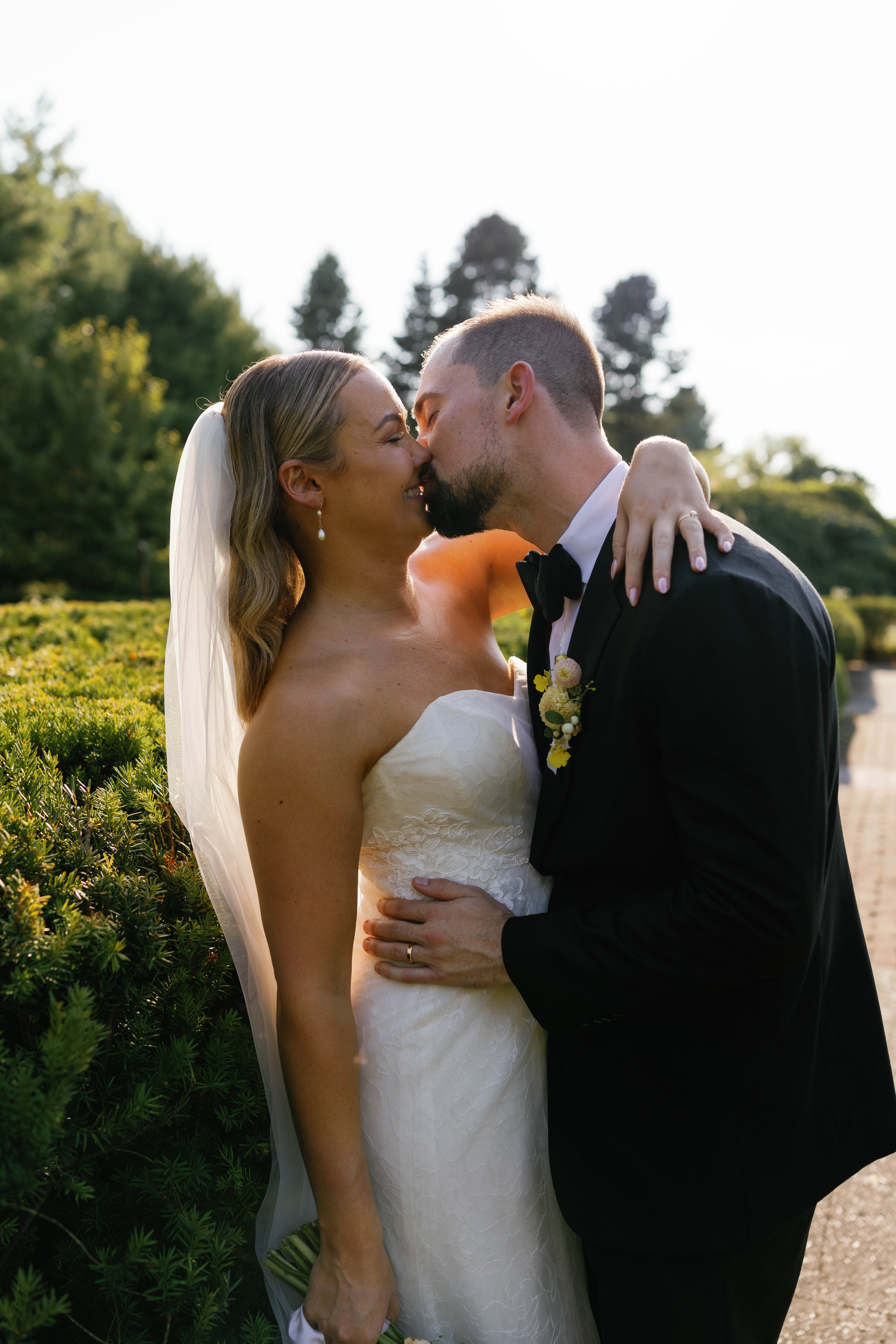 Bride leaning against garden hedge. groom pushing into bride kissing her.