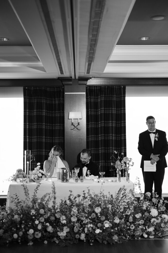 Black and white image of bride and groom at head table listening to bride's father's speech. Father standing next to head table. Bride wiping tear.