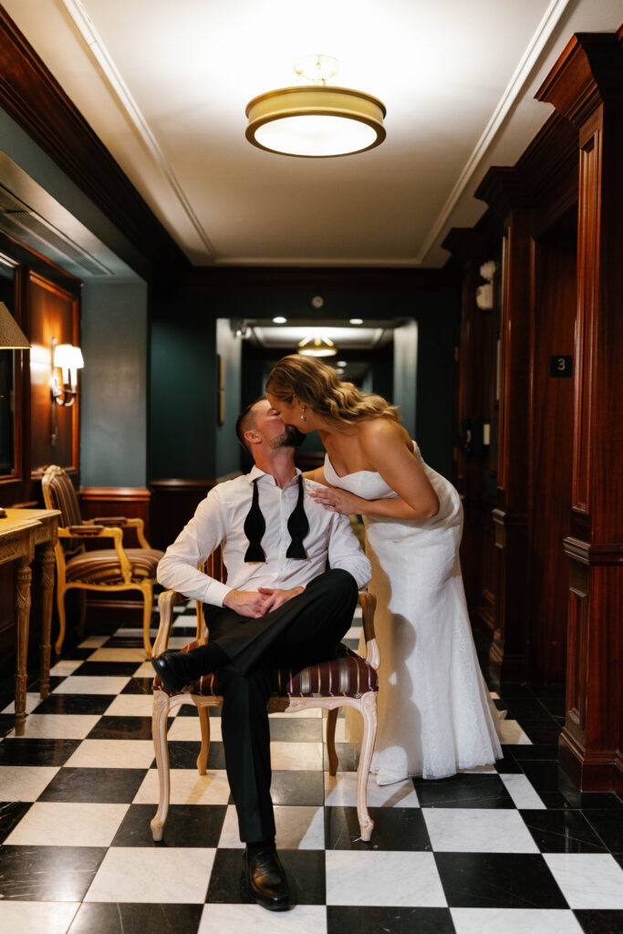 Groom sitting. Bride leaning over sharing kiss with the groom in the checkered hallway of the Graduate Hotel 