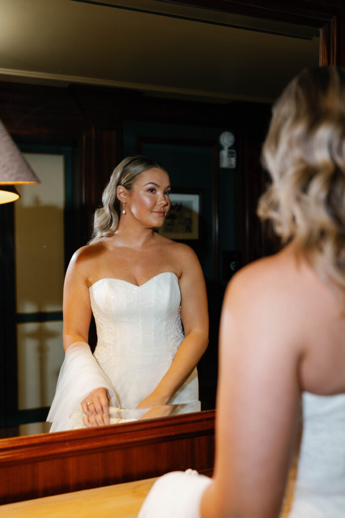 Bride looking at reflection in graduate hotel mirror.