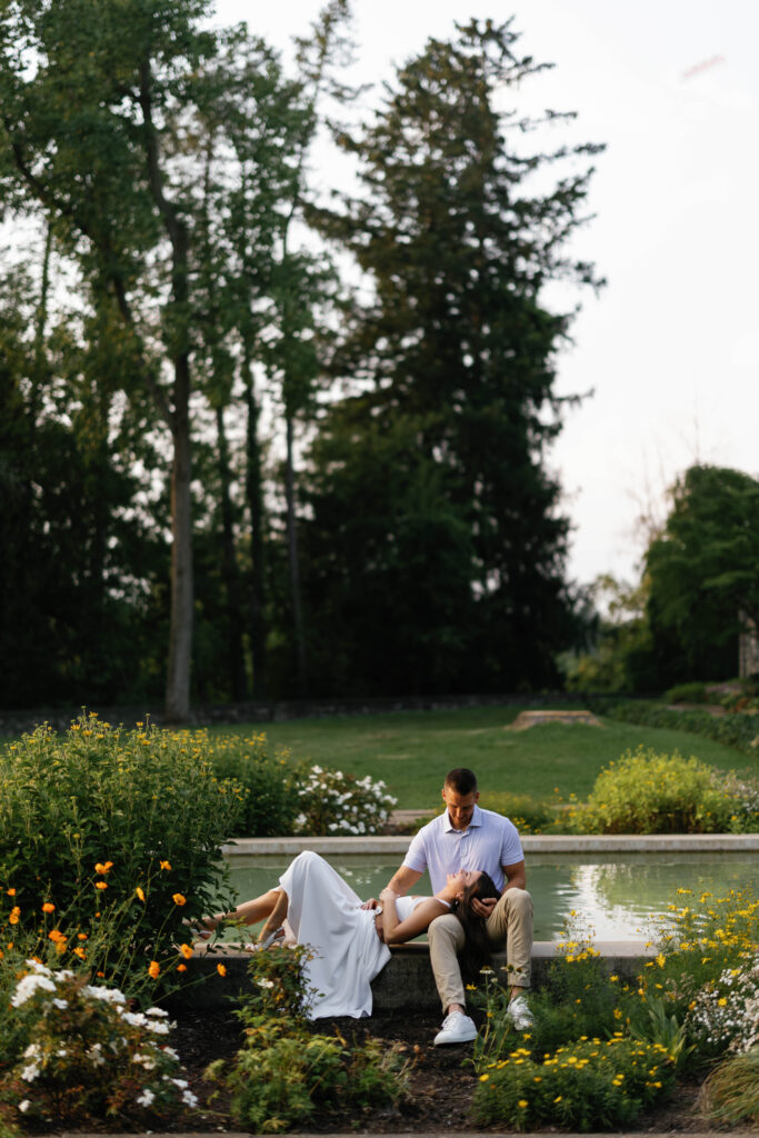 Bride to be laying in groom to be's lap on edge of reflection pool