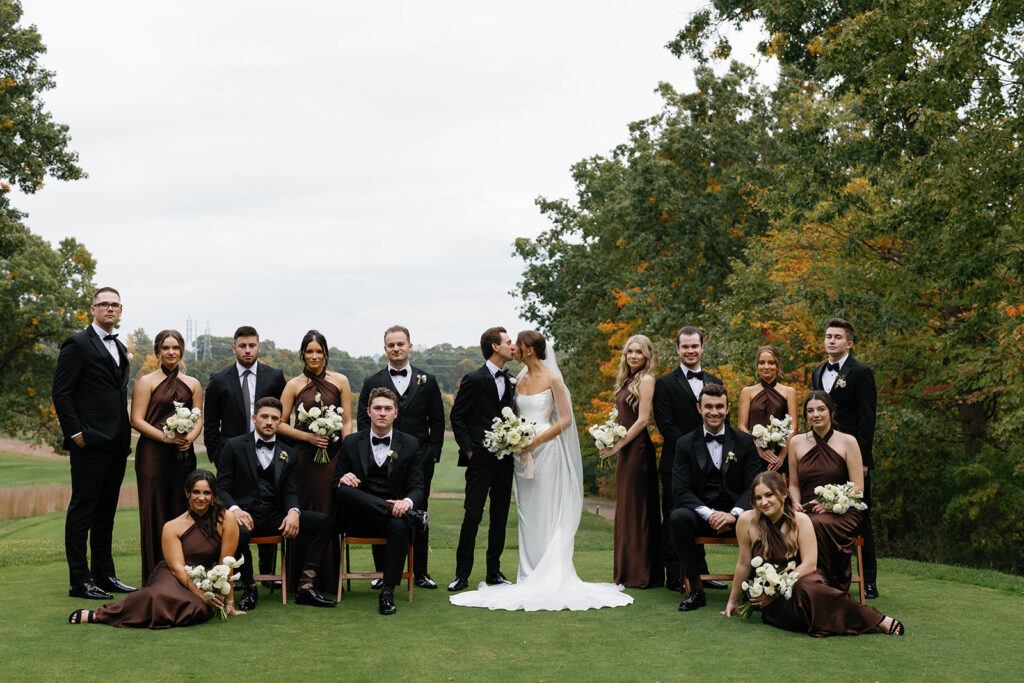 Full wedding party posed together at The Mines Golf Course with fall trees in the background during Amway Grand Plaza wedding portraits