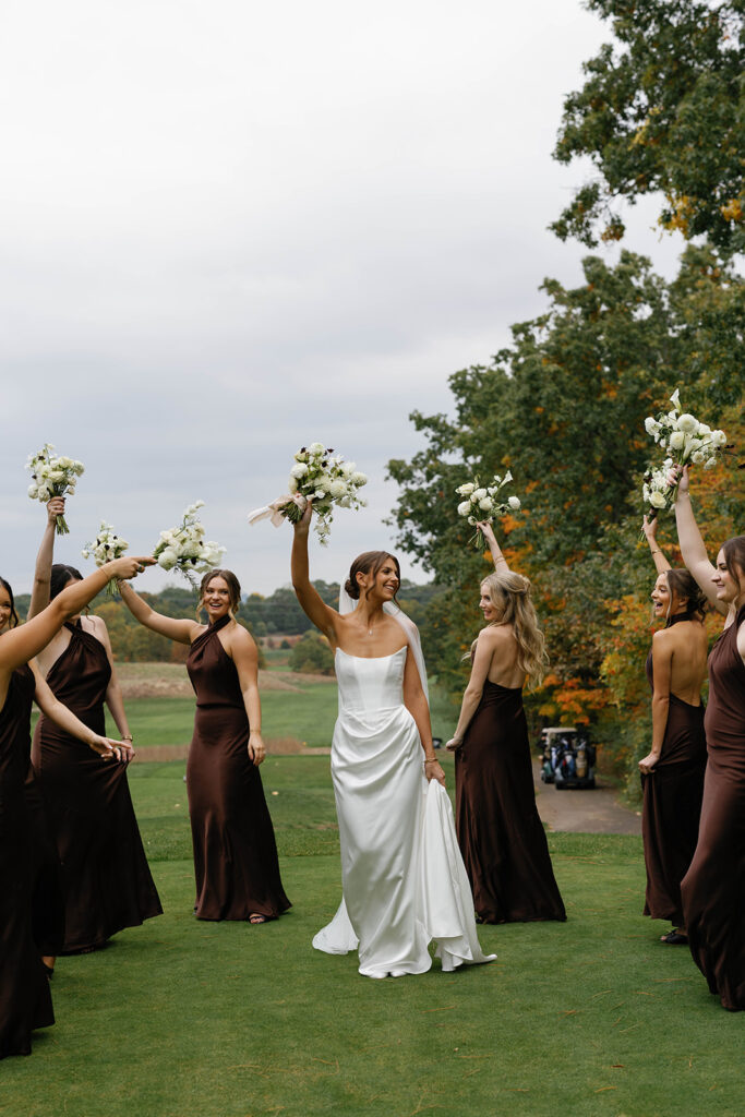 Bride and bridesmaids raising bouquets in celebration at The Mines Golf Course during Amway Grand Plaza wedding