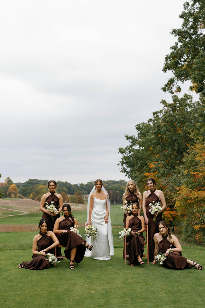 Bride with bridesmaids in brown dresses posing on golf course with autumn trees during Amway Grand Plaza wedding portraits
