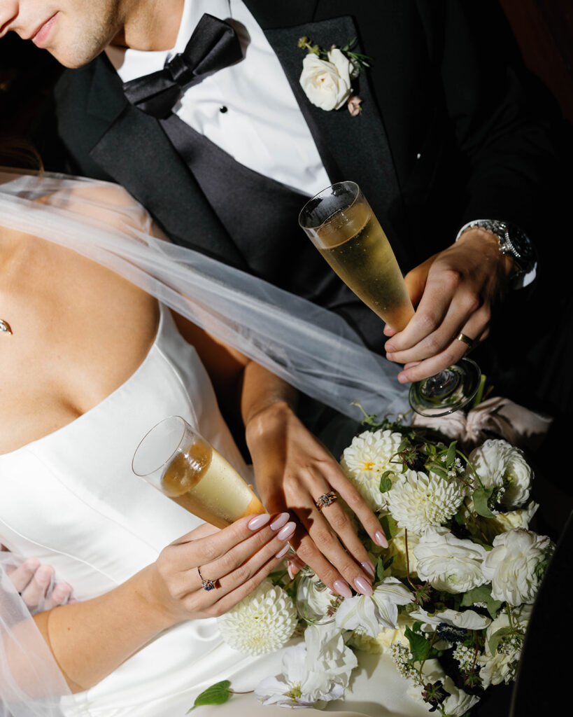Close-up of bride and groom holding champagne flutes with wedding rings and white floral bouquet at the Amway Grand Plaza