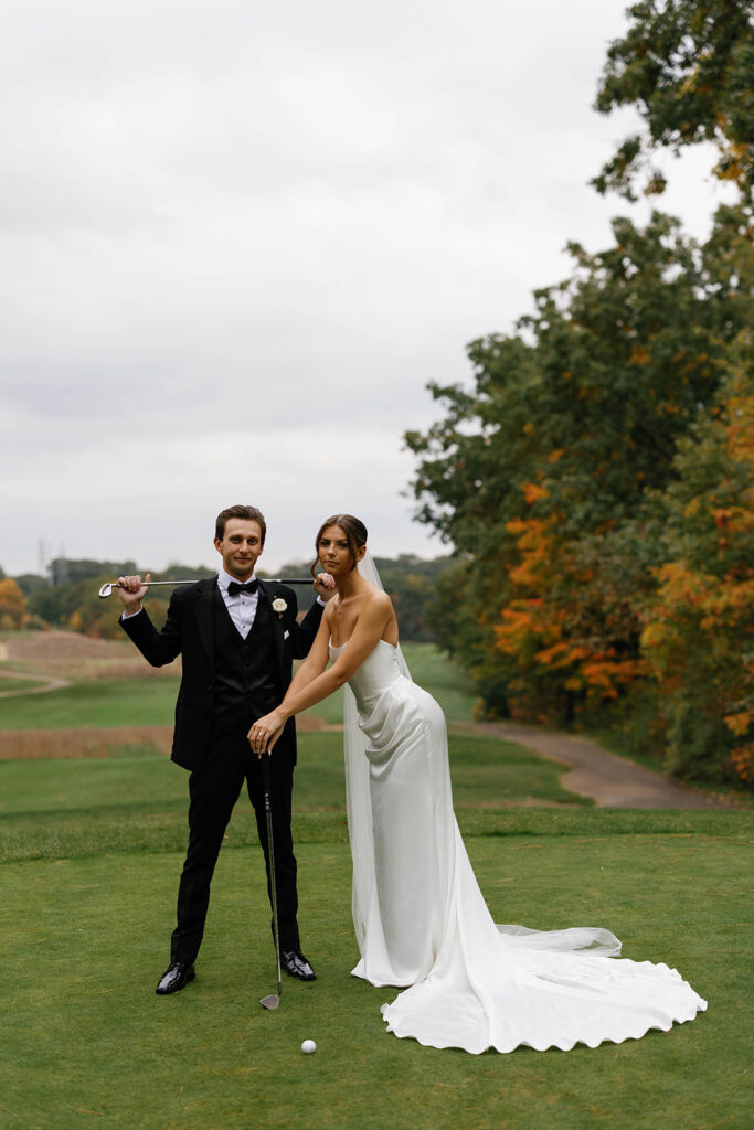 Bride and groom posing with golf clubs at The Mines Golf Course surrounded by fall colors during Amway Grand Plaza wedding