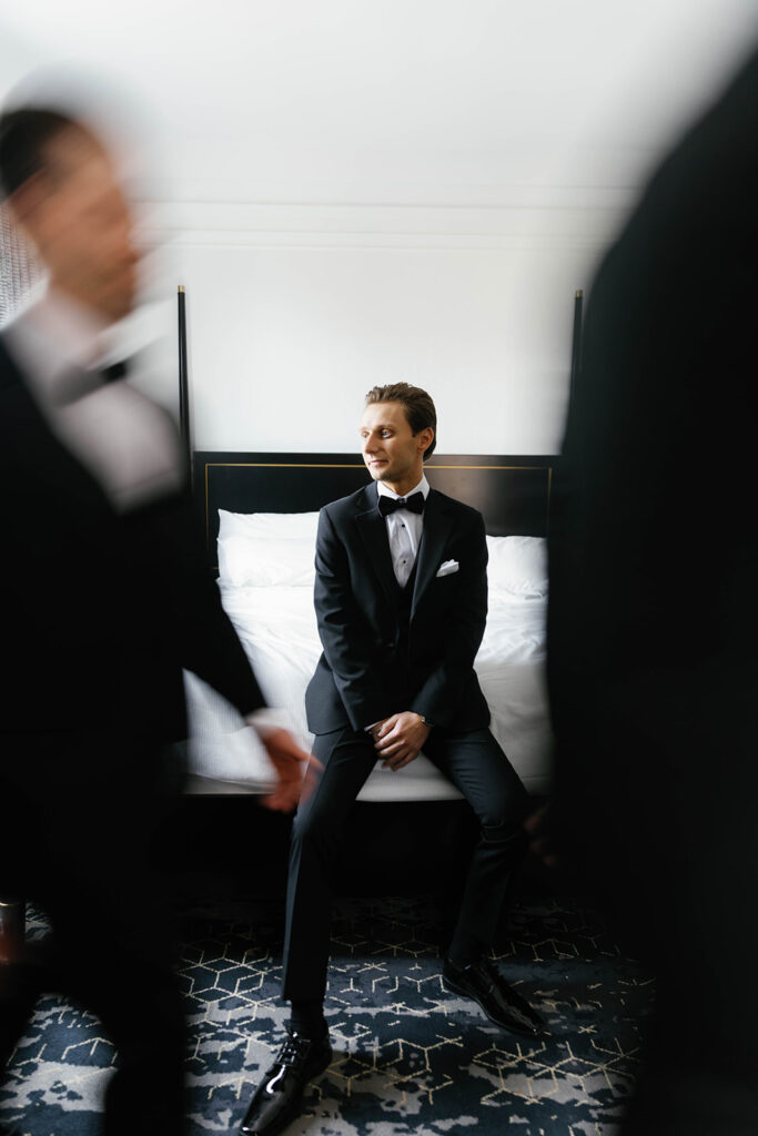 Groom seated on hotel bed in tuxedo while groomsmen move around him, editorial flash portrait at Amway Grand Plaza wedding