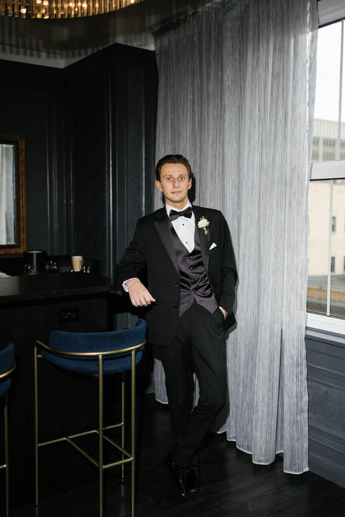 Groom in black tuxedo leaning casually against bar near window at Amway Grand Plaza wedding suite