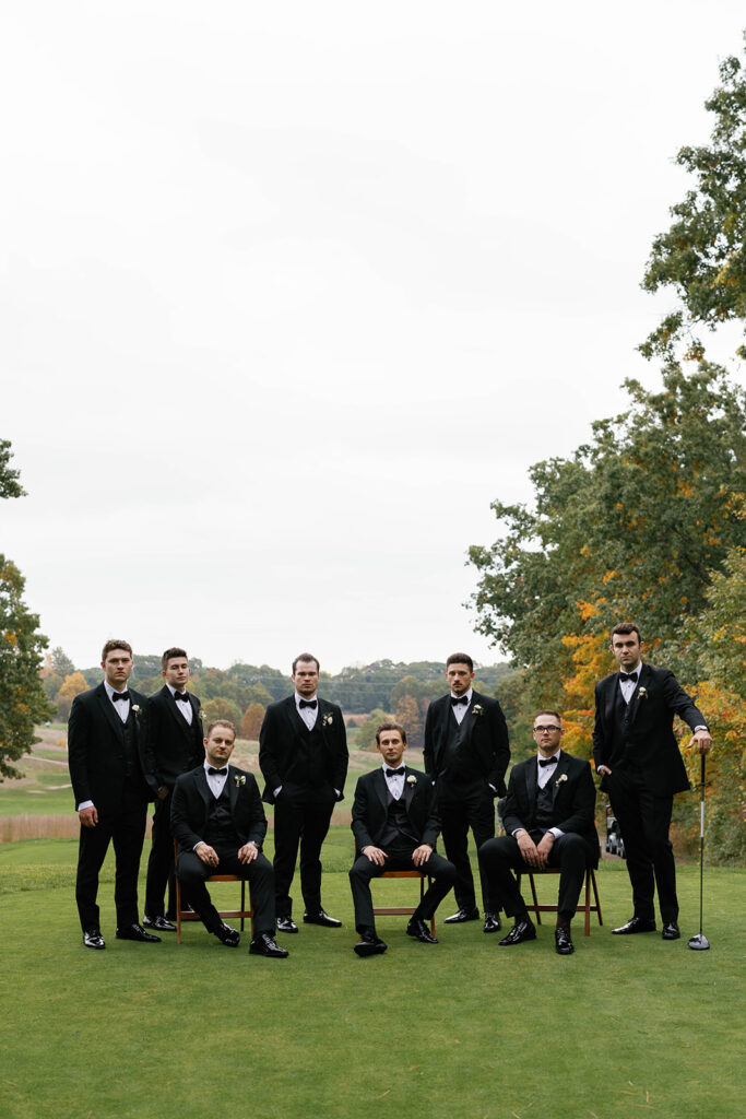 Groom and groomsmen in black tuxedos posing together at The Mines Golf Course with fall foliage during Amway Grand Plaza wedding