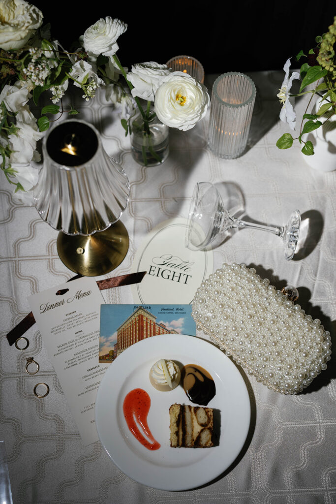Elegant flat lay of Amway Grand Plaza wedding reception table featuring dessert plate, pearl clutch, vintage postcard, and floral decor