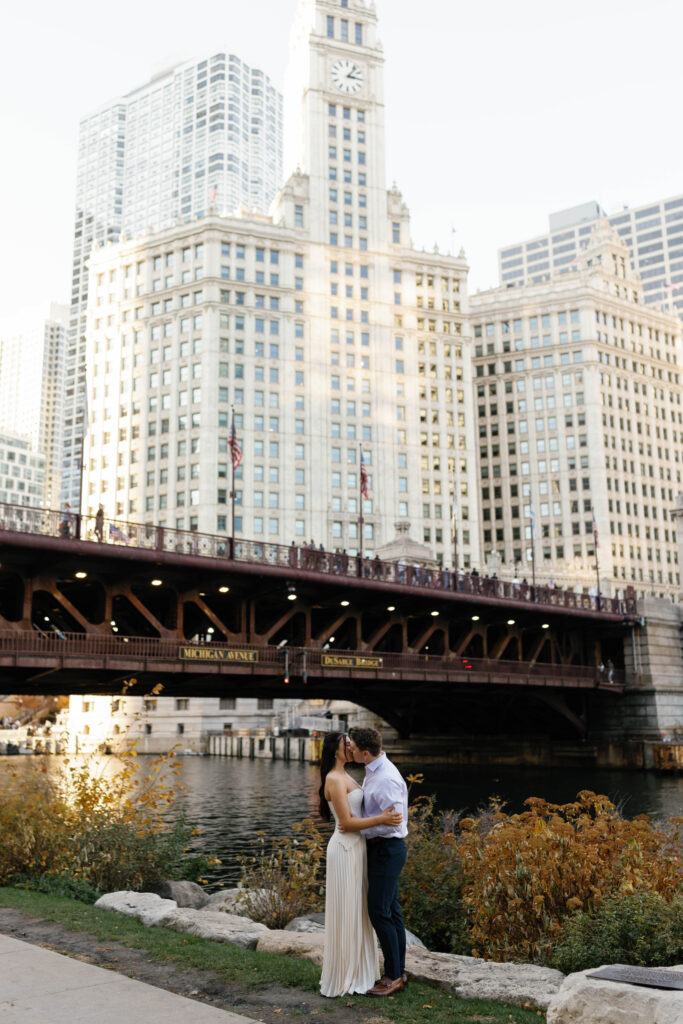 Chicago Riverwalk engagement session couple kiss at river edge with Wrigley Building skyline