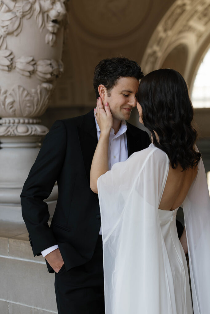 Intimate close portrait of an engaged couple at San Francisco City Hall, bride tenderly cradling her fiancé's face with her hand as they lean toward each other, ornate stone decoration in the background