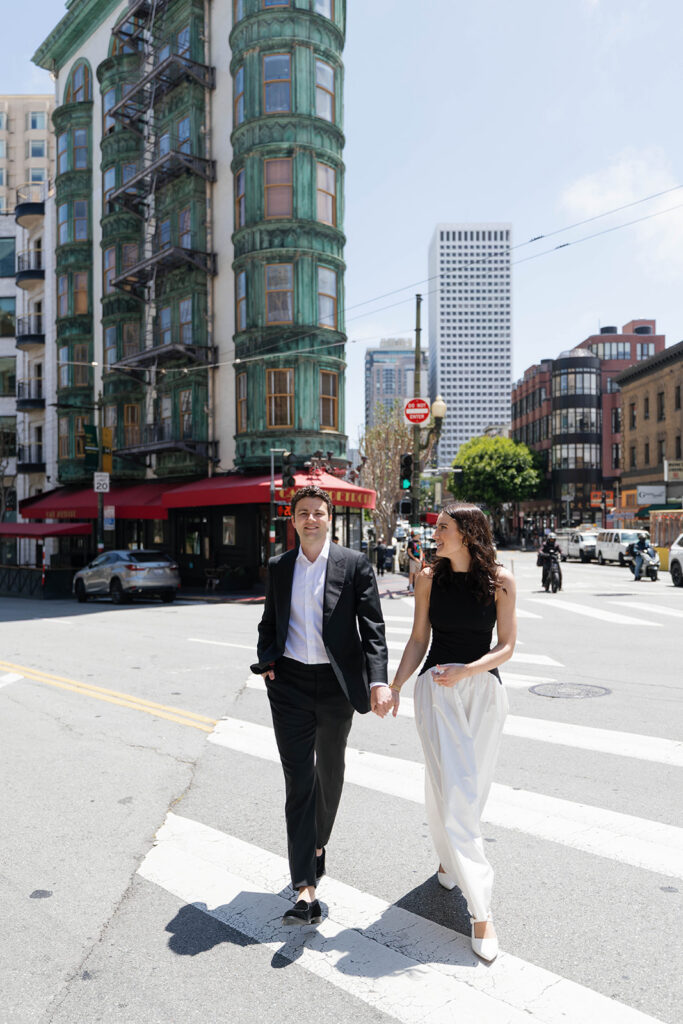 Engaged couple walking hand in hand across a sunny San Francisco crosswalk near Cafe Zoetrope, smiling at each other with the green Columbus Tower rising behind them
