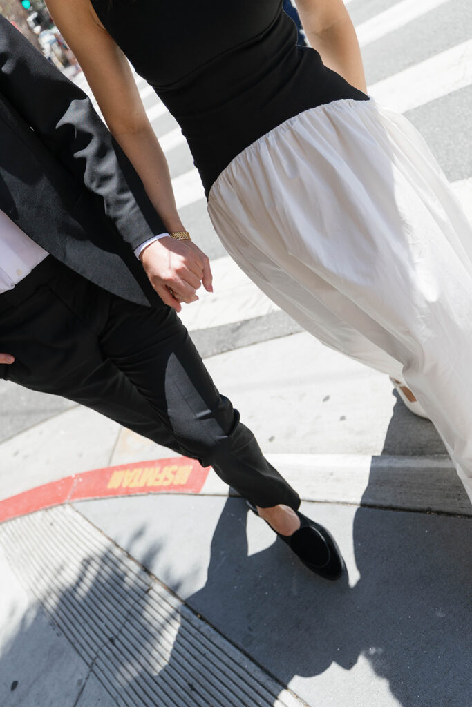 Angled detail shot of an engaged couple holding hands while walking across a San Francisco street crosswalk near North Beach, white skirt flowing as they walk
