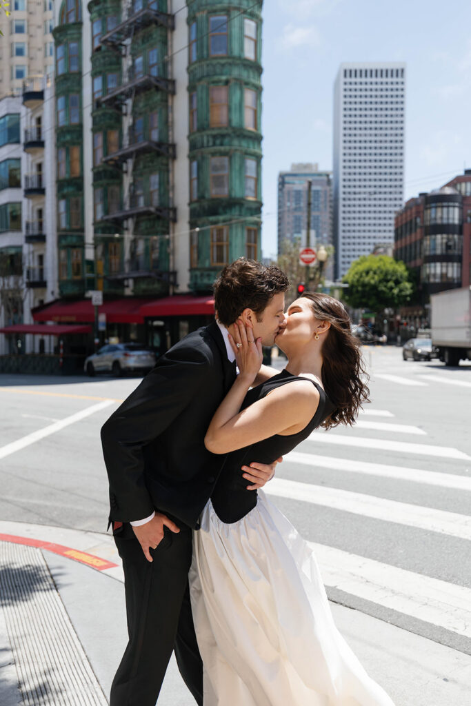 Engaged couple sharing a dip kiss in the middle of a San Francisco crosswalk with the iconic green Columbus Tower and Cafe Zoetrope visible in the background