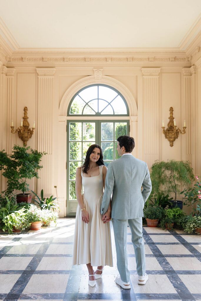 Engaged couple holding hands inside the Filoli estate conservatory, bride facing the camera in a cream midi dress as they stand on a geometric black and white marble floor beneath a grand arched window