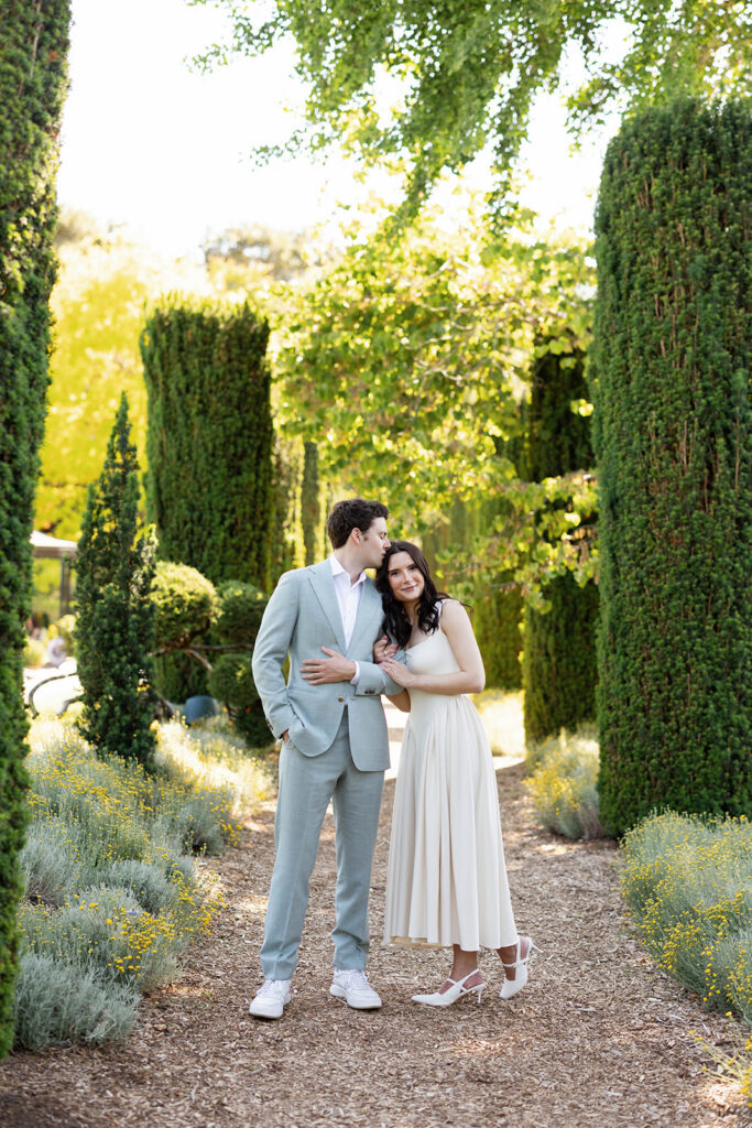Engaged couple standing together on a garden path at Filoli Gardens in Woodside California, surrounded by tall cypress trees and lavender wildflowers in golden hour light