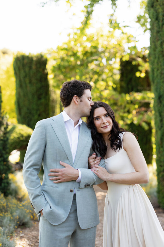 Close portrait of an engaged couple at Filoli Gardens, groom kissing his fiancée on the temple as she smiles softly at the camera with golden hour light behind them