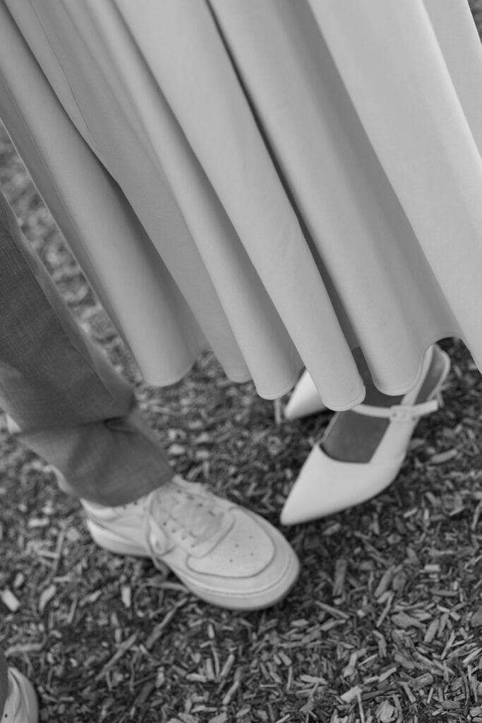 Black and white detail shot of an engaged couple's shoes at Filoli Gardens, bride in pointed heels and groom in white sneakers beneath flowing dress and suit pants