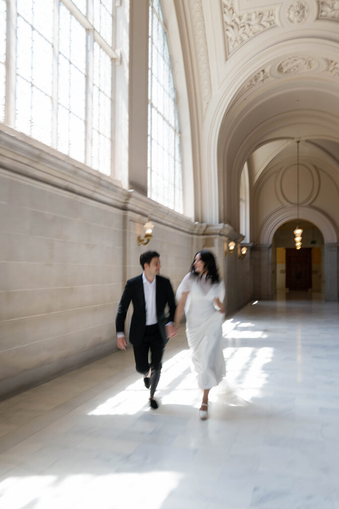 Motion blur photo of an engaged couple running and laughing together toward the camera in the sunlit marble corridor of San Francisco City Hall, warm sconce lights glowing in the background