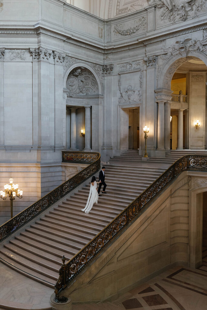 Wide overhead shot of an engaged couple ascending the grand staircase at San Francisco City Hall, iron railings and warm chandelier light framing them from above