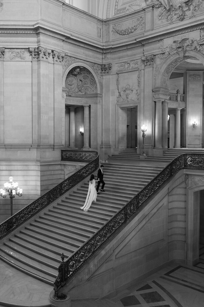 Black and white dramatic wide angle photo of an engaged couple standing small on the sweeping grand staircase at San Francisco City Hall, ornate architecture towering above them