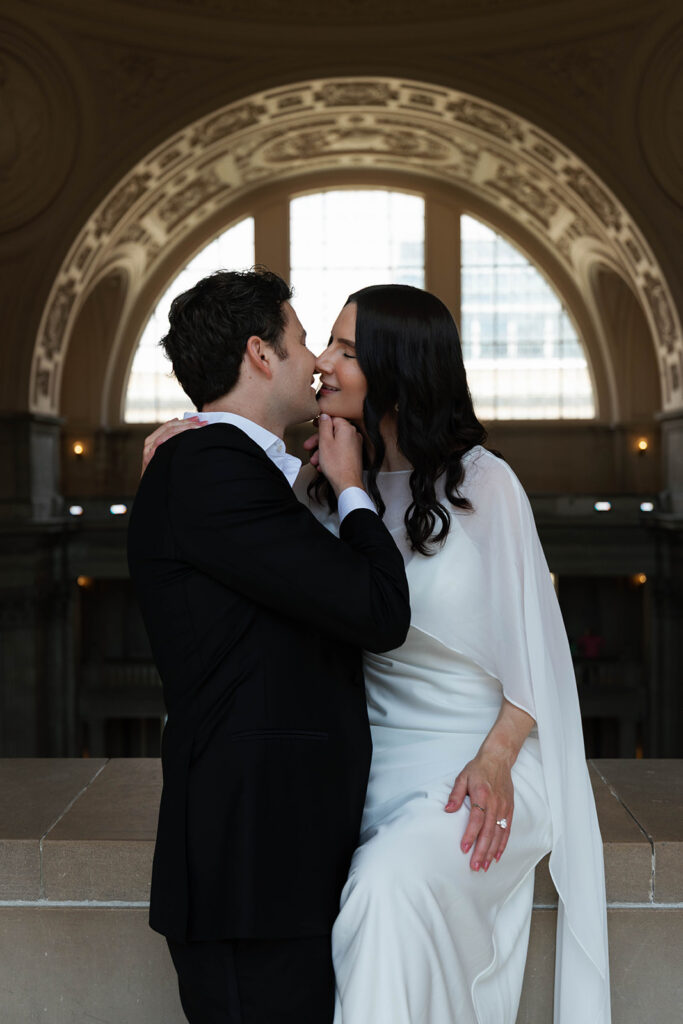 Engaged couple sharing a kiss at San Francisco City Hall, beautifully backlit by a large arched window that wraps them in soft golden light