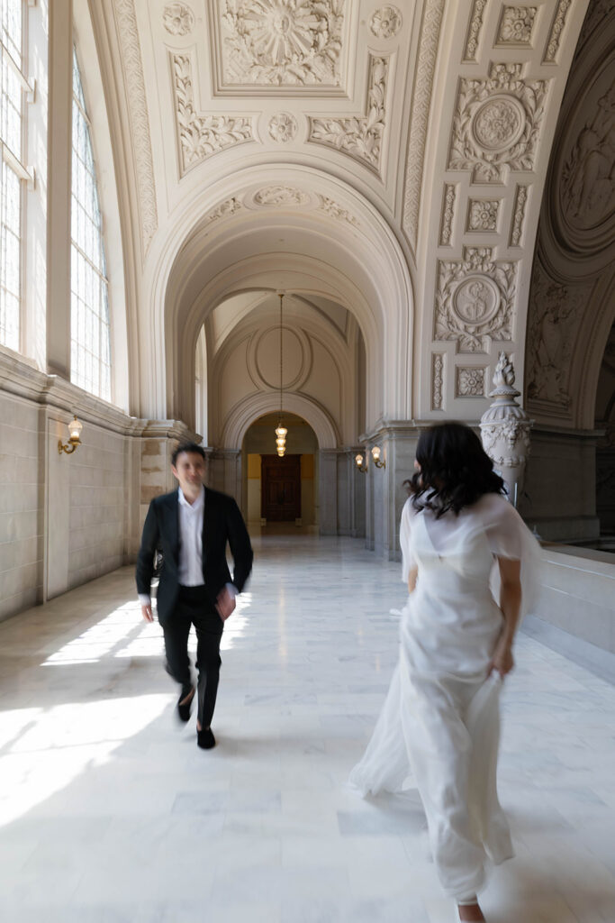 Motion blur photo of a groom running toward his fiancée in the grand marble corridor of San Francisco City Hall, intricate plasterwork ceiling and arched doorways stretching behind them