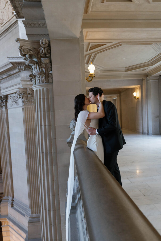 Engaged couple sharing a kiss while leaning against the stone railing at San Francisco City Hall, tall Corinthian columns and a warm gold wall sconce framing the moment