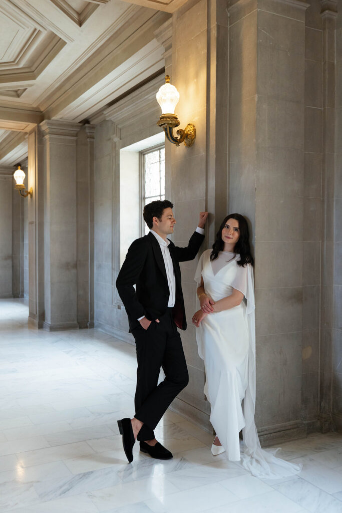 Engaged couple leaning against a stone column in a warmly lit hallway at San Francisco City Hall, brass wall sconce glowing above them as they the groom looks at the bride and the bride looks at the camera