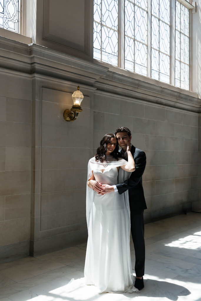 Engaged couple embracing next to a stone wall at San Francisco City Hall, soft light filtering through leaded glass windows with a brass wall sconce above them