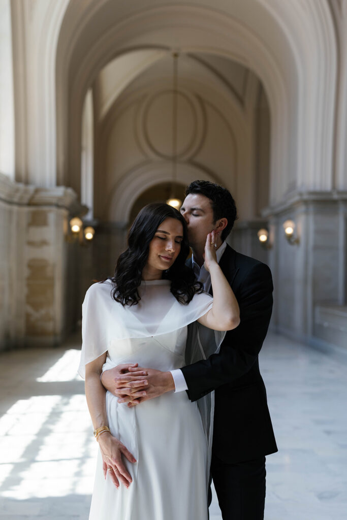 Intimate portrait of an engaged couple at San Francisco City Hall, groom gently cupping his fiancée's face as she closes her eyes, soft arched hallway and warm sconce light behind them