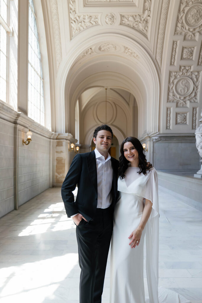 Engaged couple smiling together at the camera in a sun-drenched marble hallway at San Francisco City Hall, ornate arched ceilings and warm sconce lighting behind them